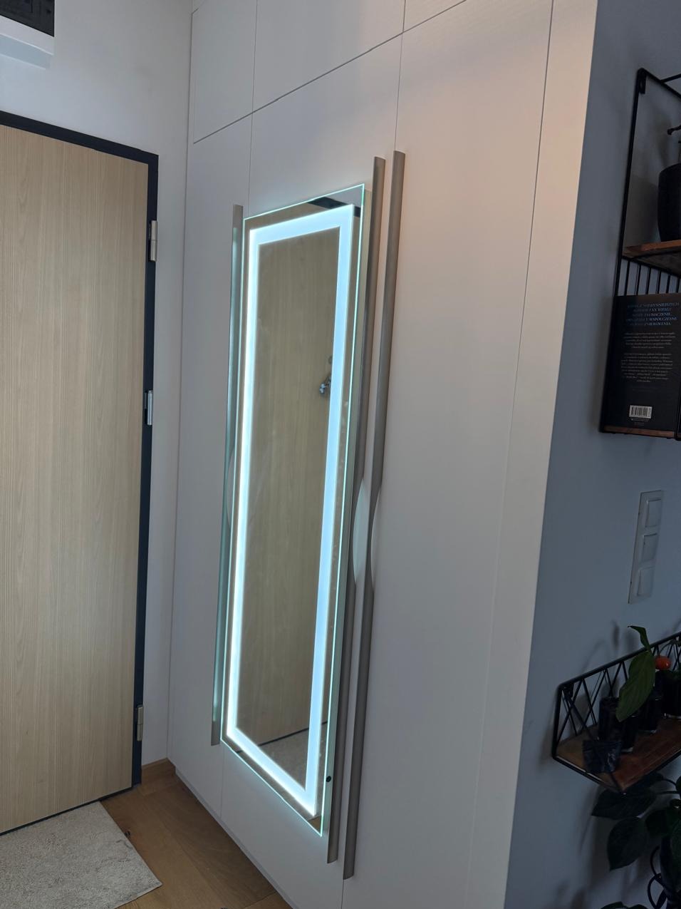 Biała szafa wnękowa z lustrem podświetlanym LED, widok z boku, drzwi drewniane w tle. Minimalistyczny design, nowoczesne oświetlenie.