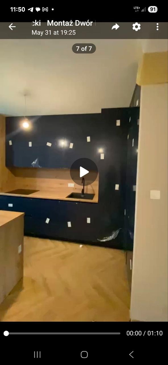 Ciemna zabudowa kuchenna z drewnianym blatem, zabezpieczona folią i taśmą malarską, na podłodze parkiet. Widok na fragment wnętrza w trakcie montażu.