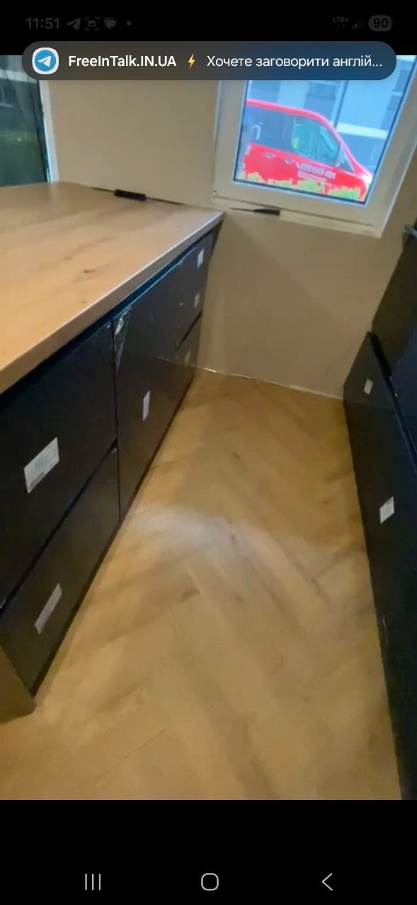 Ciemne szafki z jasnym blatem pod oknem na poddaszu, parkiet w jodełkę. Widok na czerwony samochód z okna.