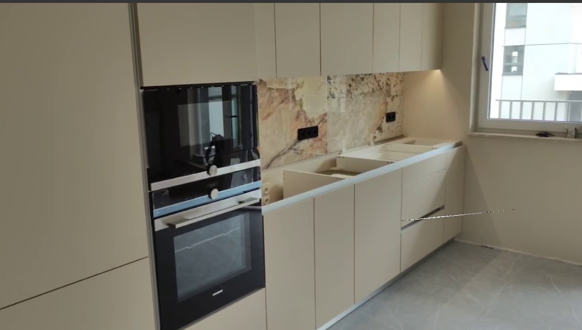 Beżowa zabudowa kuchenna z piekarnikiem i płytą grzewczą, marmurowy backsplash, widok na okno i fragment budynku.