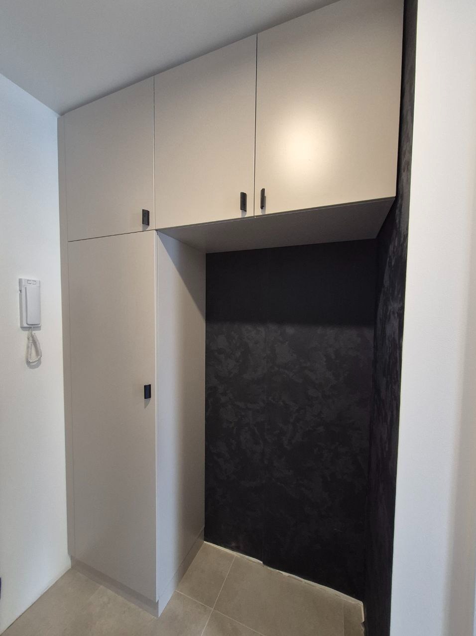 Jasnoszara szafa wnękowa z czarnymi uchwytami, w przedpokoju z ciemną ścianą o fakturze. Minimalistyczny design, proste linie.