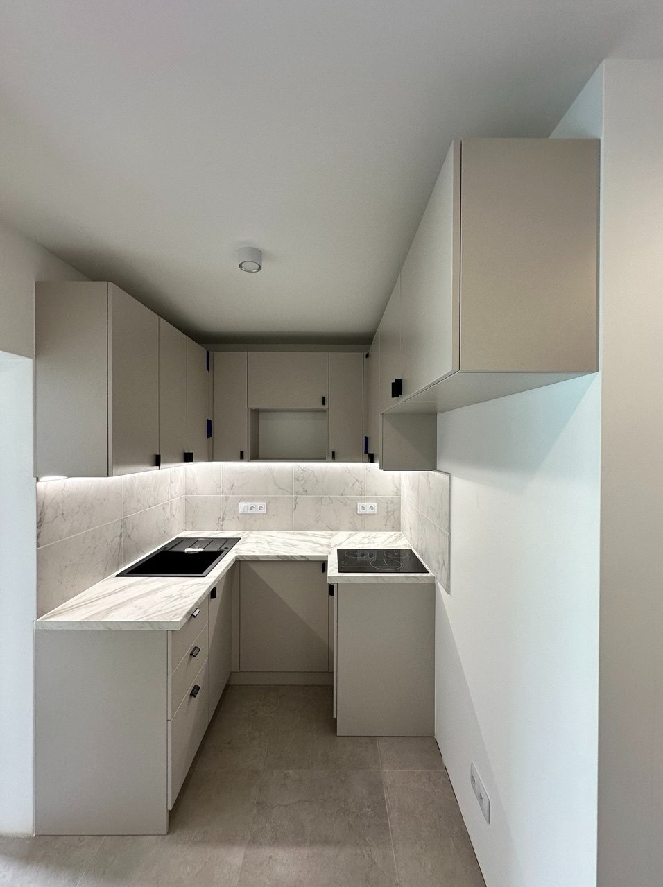 Kompaktowa kuchnia w kształcie litery U z marmurowym blatem i podświetleniem LED. Minimalistyczne, jasne fronty szafek i czarne, smukłe uchwyty. Płyta indukcyjna i zlewozmywak w czarnym kolorze.