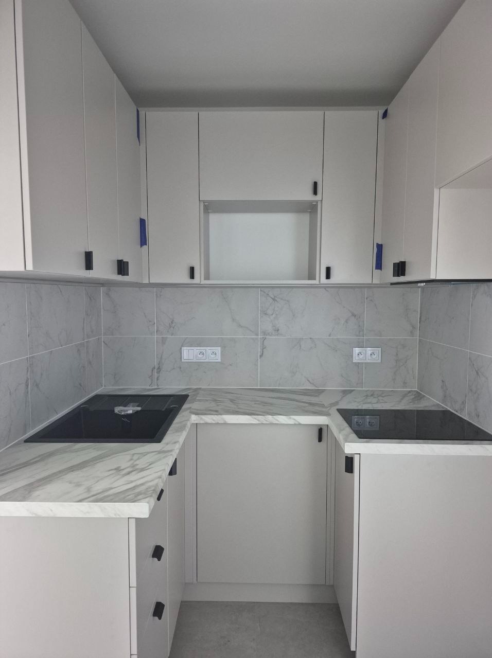 Nowoczesna, jasna kuchnia z marmurowym blatem i płytkami, czarnymi uchwytami. Widoczne gniazdka elektryczne i płyta indukcyjna. Minimalistyczny design.