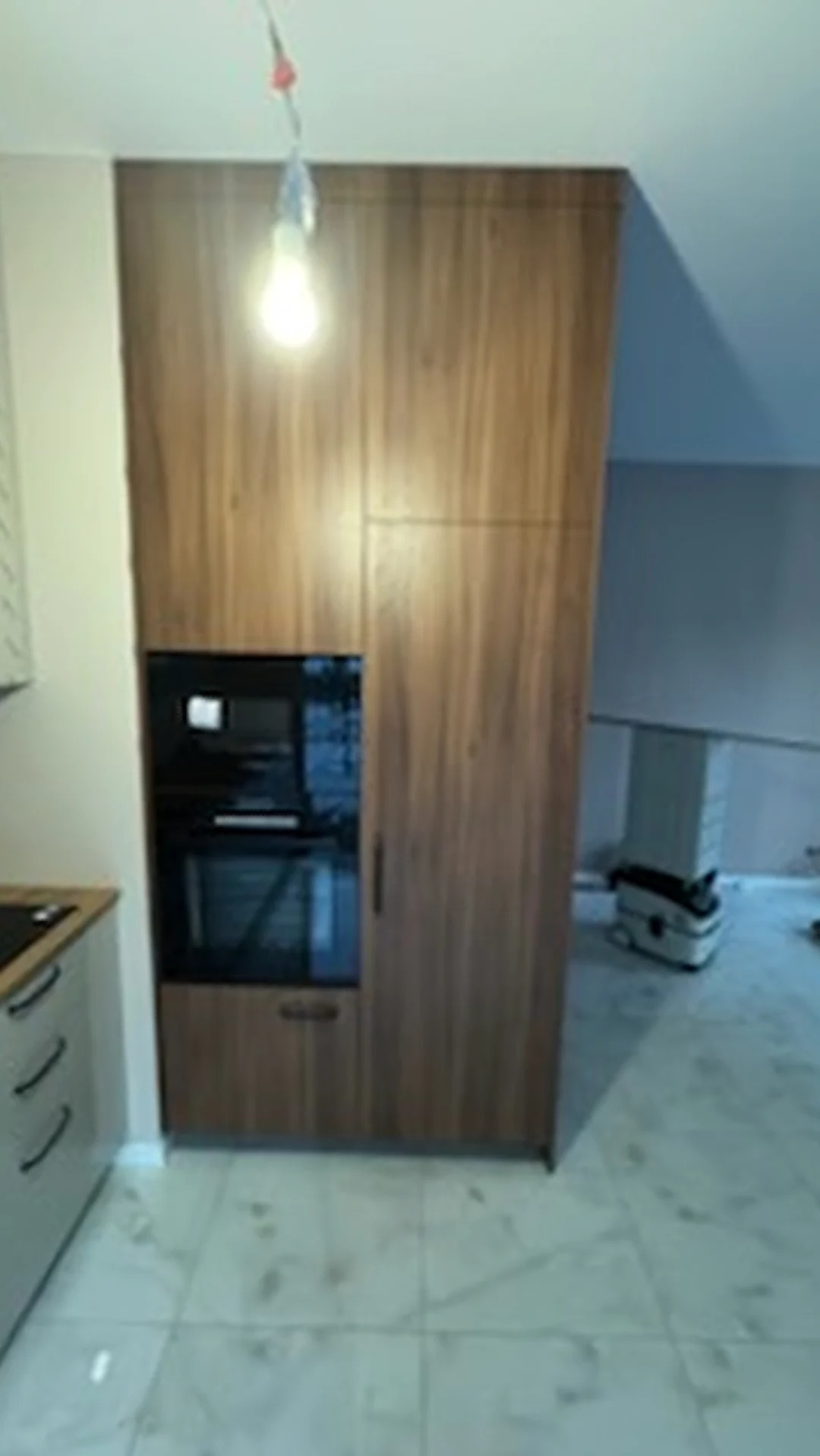 Wysoka zabudowa kuchenna z forniru orzechowego z wbudowanym piekarnikiem i mikrofalówką, na tle marmurowej podłogi i odkurzacza przemysłowego.