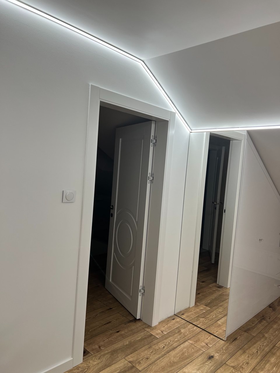 Białe drzwi wewnętrzne z dekoracyjnym frezem, częściowo otwarte, na tle drewnianej podłogi i białych ścian z oświetleniem LED. Widok z dołu, podkreślający wysokość pomieszczenia.