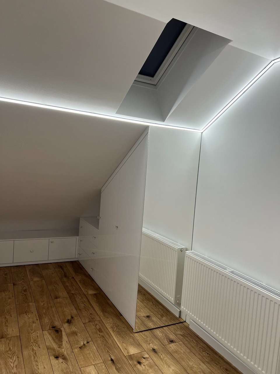 Biała szafa wnękowa z lustrzanymi drzwiami na poddaszu, drewniana podłoga, okno dachowe i oświetlenie LED. Minimalistyczny design.