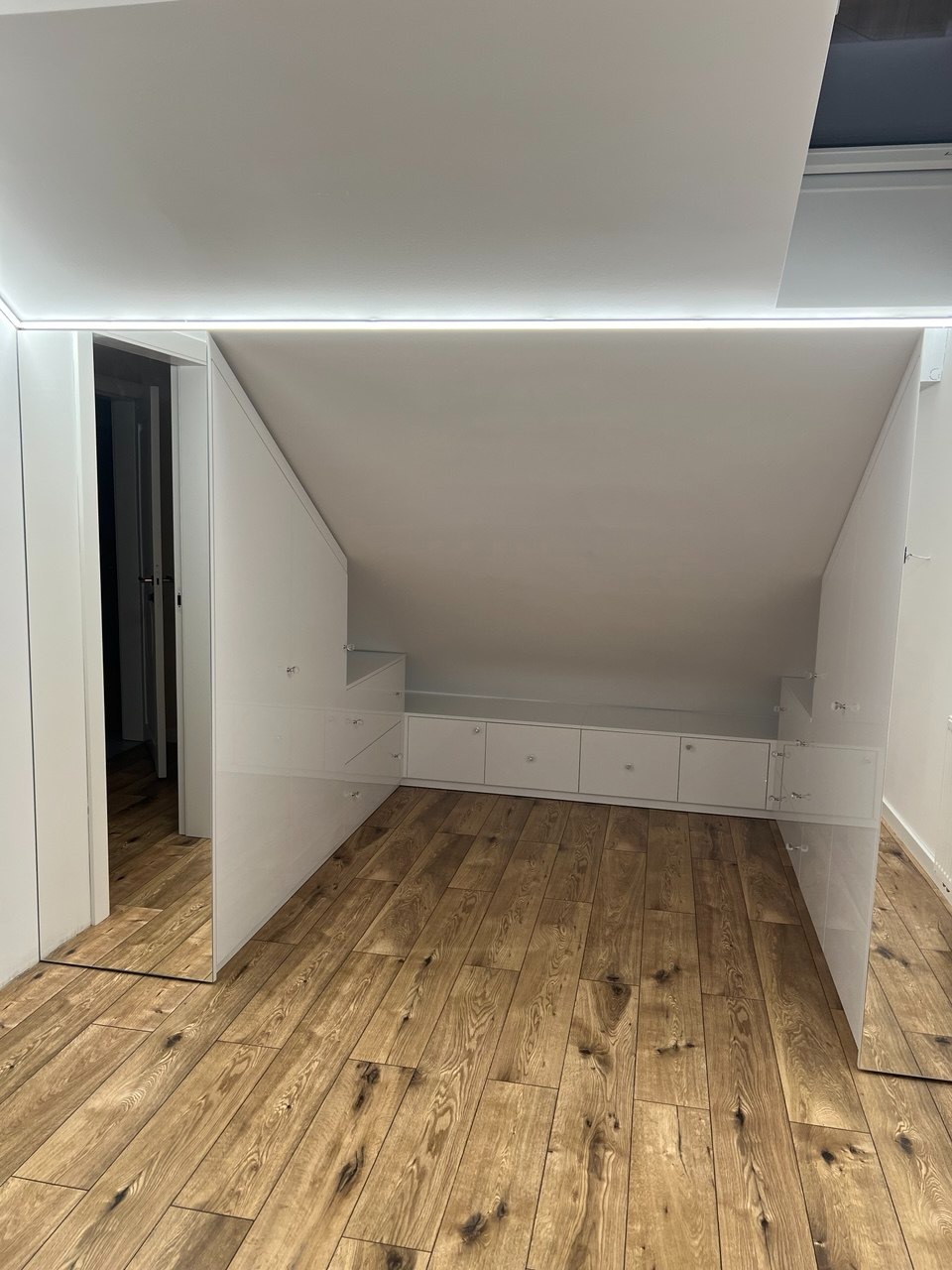 Biała zabudowa pod skosem na poddaszu z szafkami i drzwiami, drewniana podłoga, oświetlenie LED. Minimalistyczny design, jasne wnętrze.