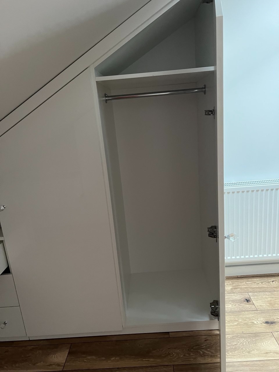 Biała szafa wnękowa z otwartymi drzwiami, widoczny drążek na ubrania i półka, dopasowana do skosu dachu, drewniana podłoga, minimalistyczny design.