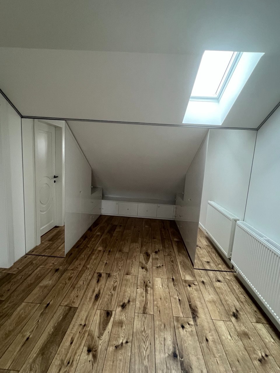 Biała zabudowa meblowa pod skosem na poddaszu z drewnianą podłogą w Warszawie. Widoczne szafki i drzwi. Okno dachowe wpuszcza światło.