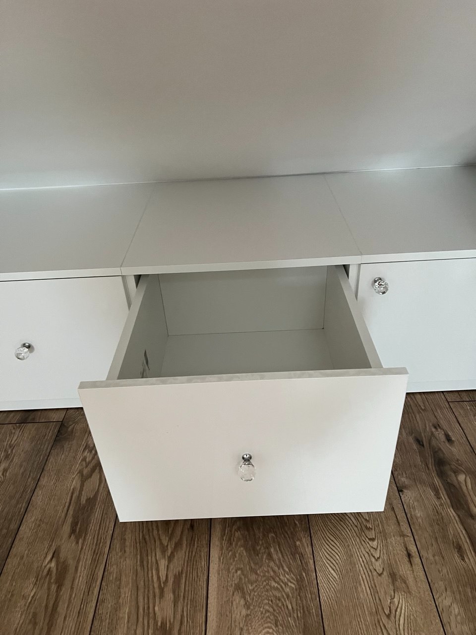 Biała szafka z szufladami na drewnianej podłodze. Jedna szuflada jest otwarta, ukazując wnętrze. Gałki szuflad są kryształowe.