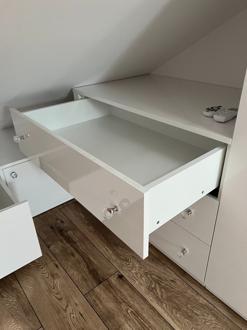 Biała szafa wnękowa z otwartą szufladą i kryształowymi gałkami, dopasowana do skosu poddasza. Na blacie leżą dwa piloty. Parkiet na podłodze.