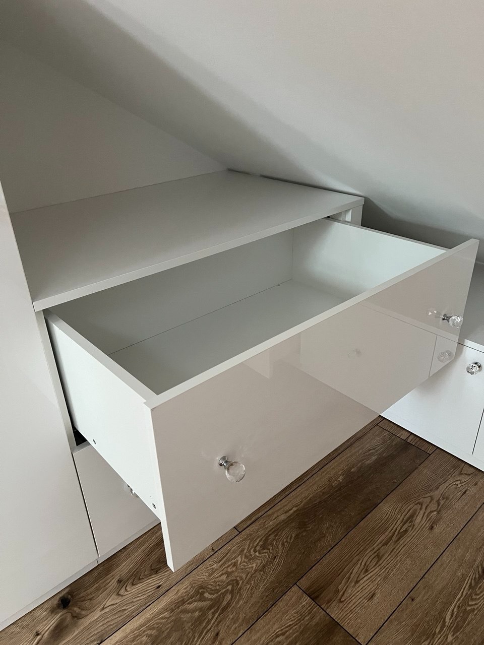 Biała szafa wnękowa z otwartą szufladą, widoczne drewniane panele podłogowe. Szuflada z błyszczącym frontem i kryształowymi uchwytami. Zabudowa pod skosem.