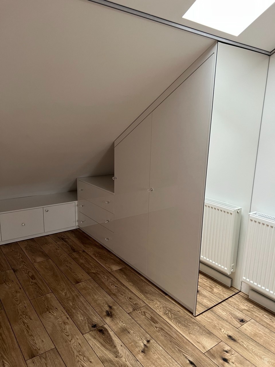 Biała szafa wnękowa na poddaszu ze schodkową zabudową i lustrem, drewniana podłoga, widoczne grzejniki i okno dachowe. Minimalistyczny design.