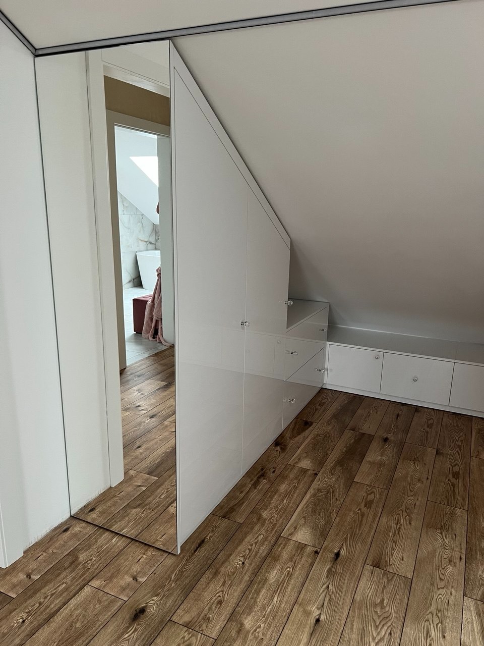 Biała szafa wnękowa z lustrzanym frontem, dopasowana do skosu poddasza. Widoczna drewniana podłoga i fragment łazienki w odbiciu lustra.