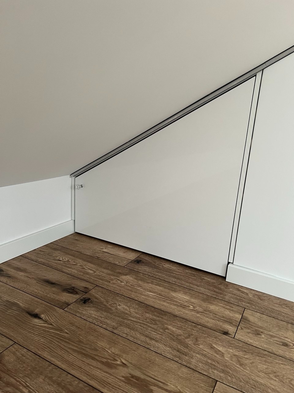 Biała szafa wnękowa pod skosem na poddaszu z drewnianą podłogą. Minimalistyczny design, widoczne detale wykończenia i kąt ujęcia podkreślający specyfikę zabudowy.