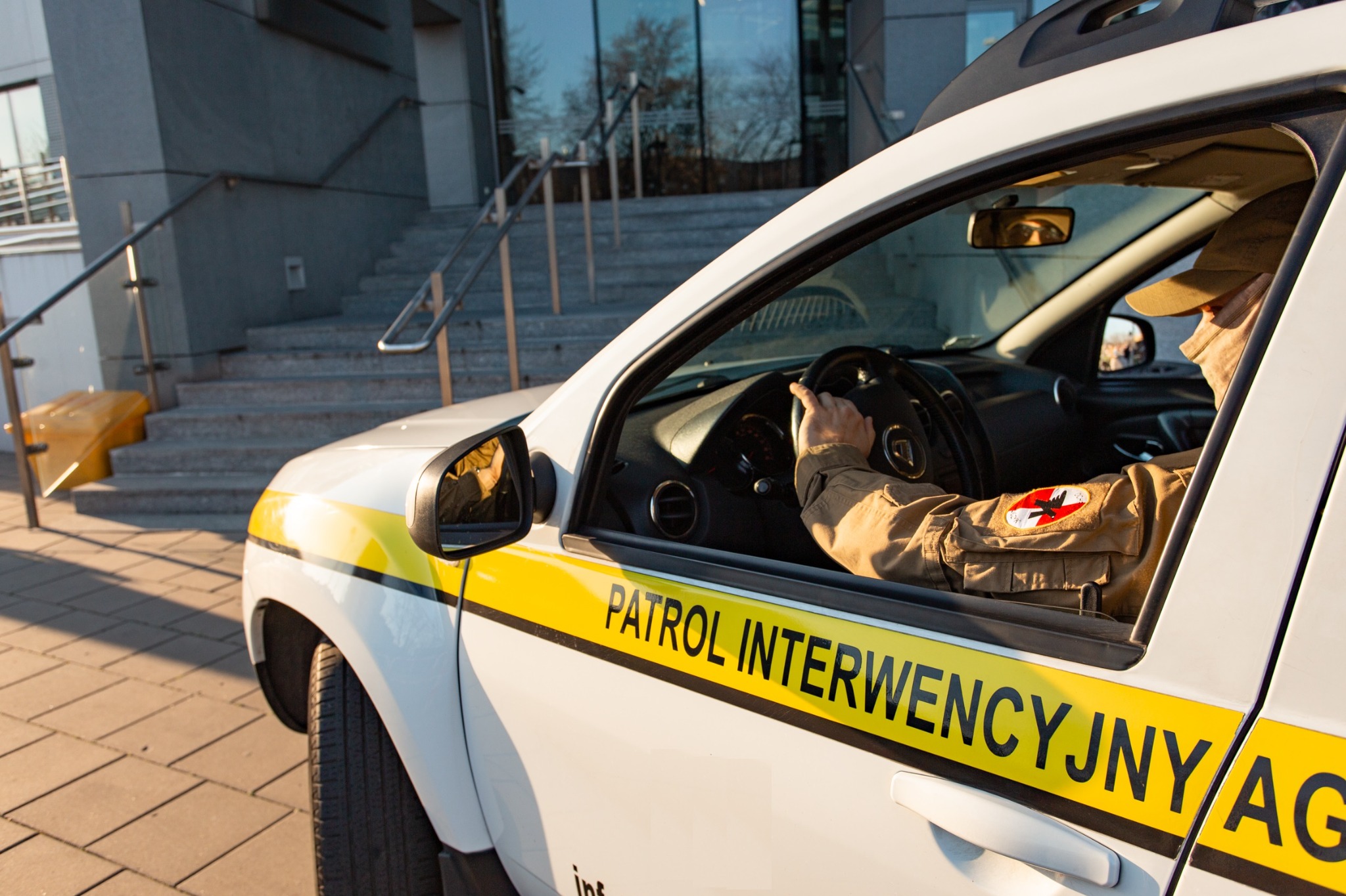 Samochód patrolowy z napisem 'PATROL INTERWENCYJNY' na tle budynku z schodami, wewnątrz kierowca w uniformie z naszywką z symbolem.