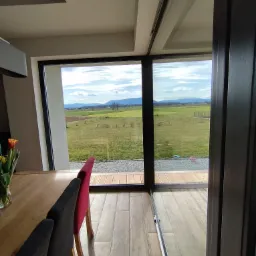 Wnętrze domu z dużym oknem tarasowym, widok na zielony trawnik z elementami infrastruktury sportowej i odległe góry, drewniana podłoga i krzesła w jadalni.