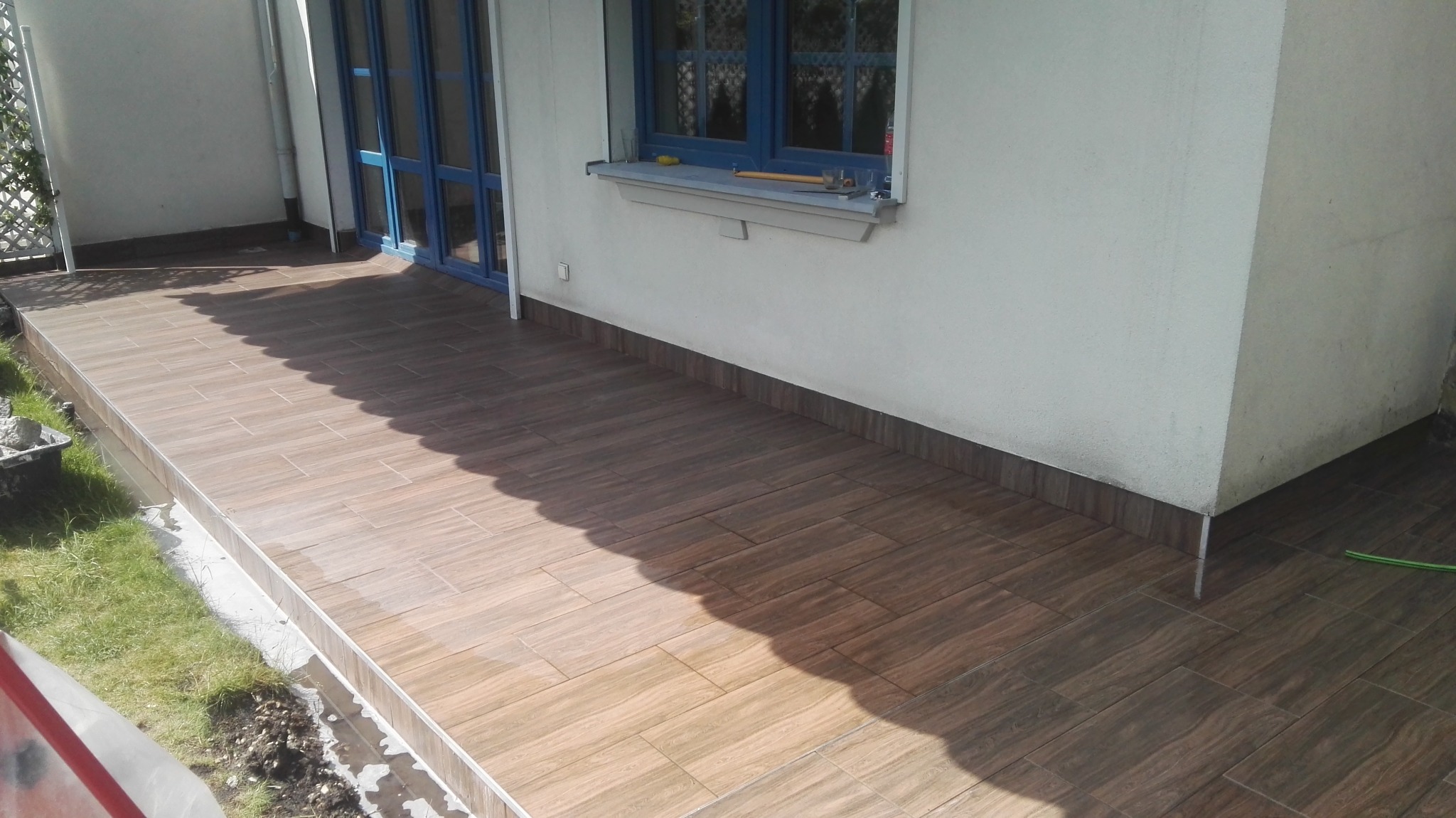 Wyłożony brązowymi płytkami taras przylegający do białej ściany budynku z niebieskimi drzwiami i oknem, na parapecie okna leżą narzędzia.