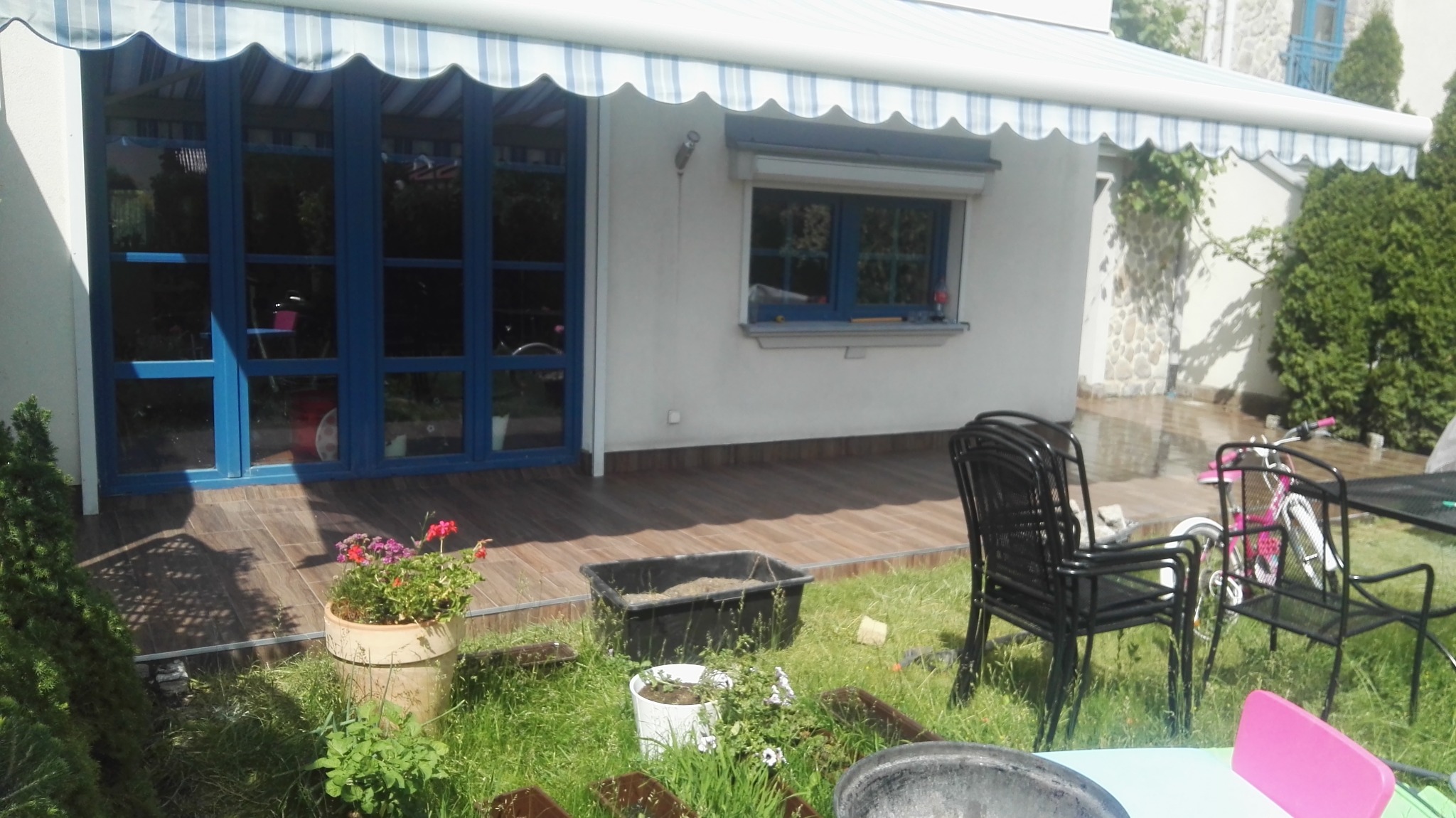 Zewnętrzny widok domu z zabudowanym balkonem, niebieska stolarka okienna i drzwiowa, drewniana podłoga na balkonie, markiza w biało-niebieskie paski, meble ogrodowe i rośliny w donicach.