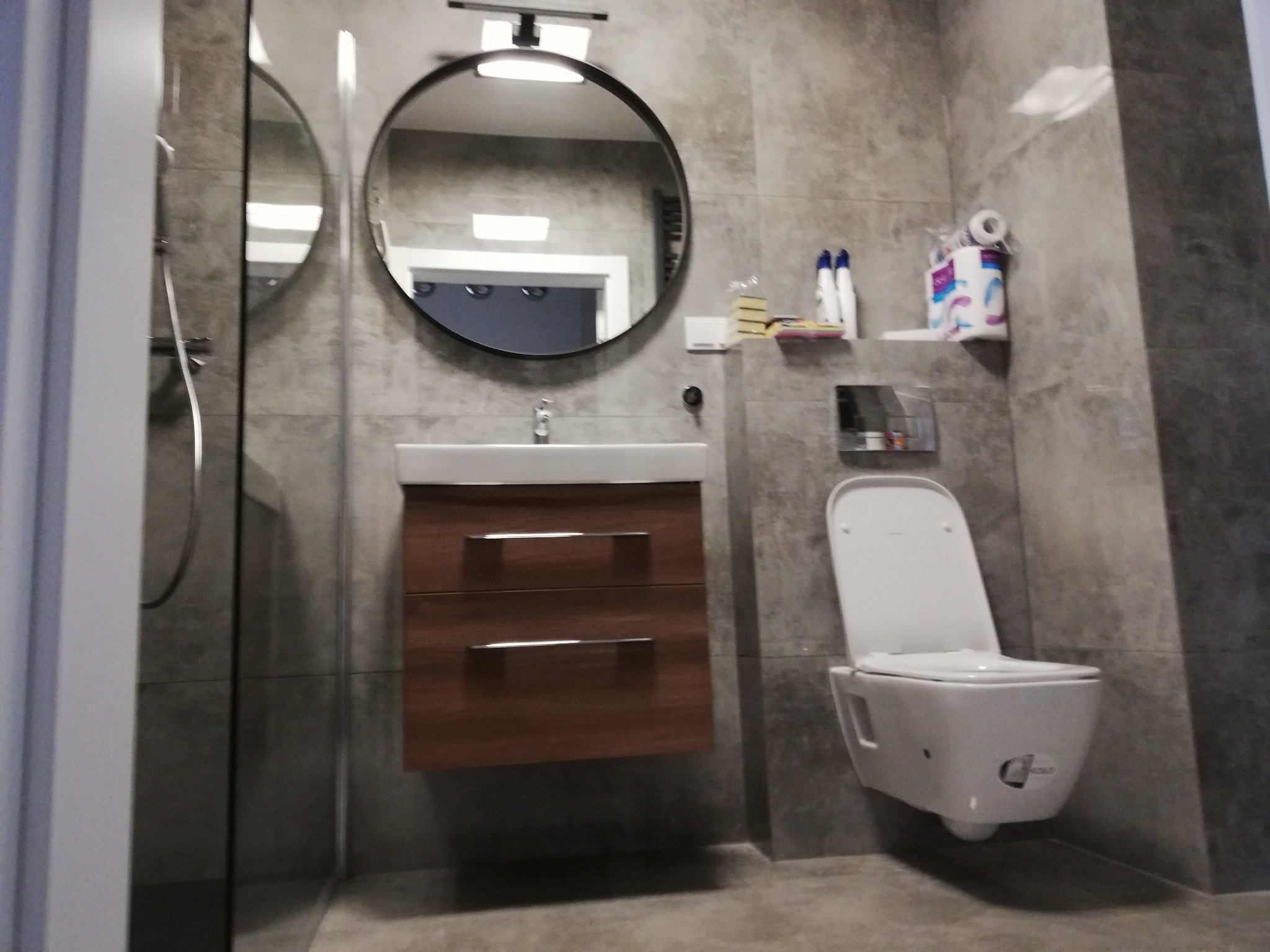 Nowoczesna łazienka z szarymi płytkami, okrągłym lustrem z podświetleniem, wiszącą szafką pod umywalką z drewnianymi szufladami i podwieszanym WC.