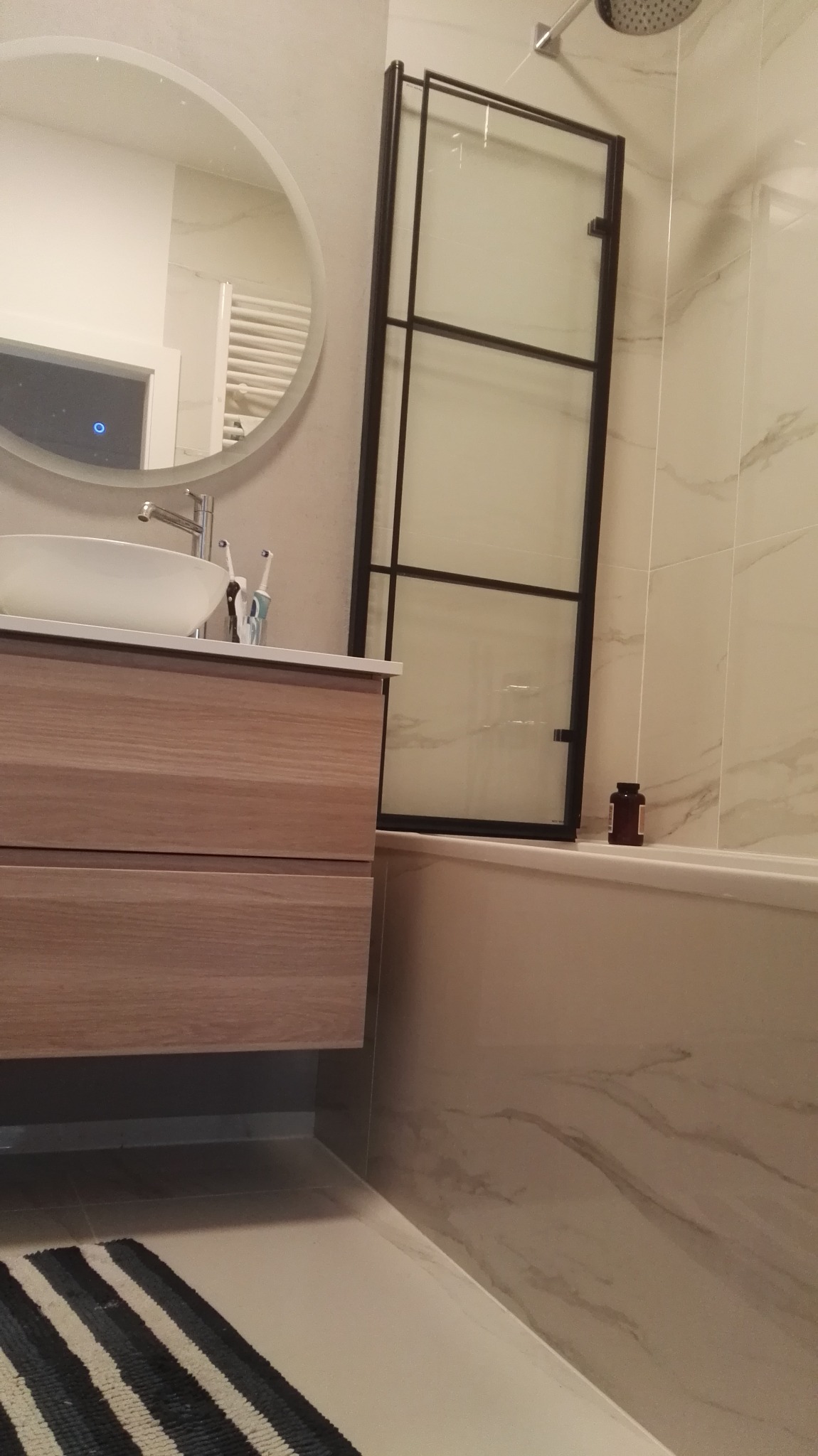 Nowoczesna łazienka z umywalką nablatową na drewnianej szafce, owalnym lustrem z podświetleniem LED, kabiną prysznicową z czarnymi ramami i marmurową okładziną ścienną.