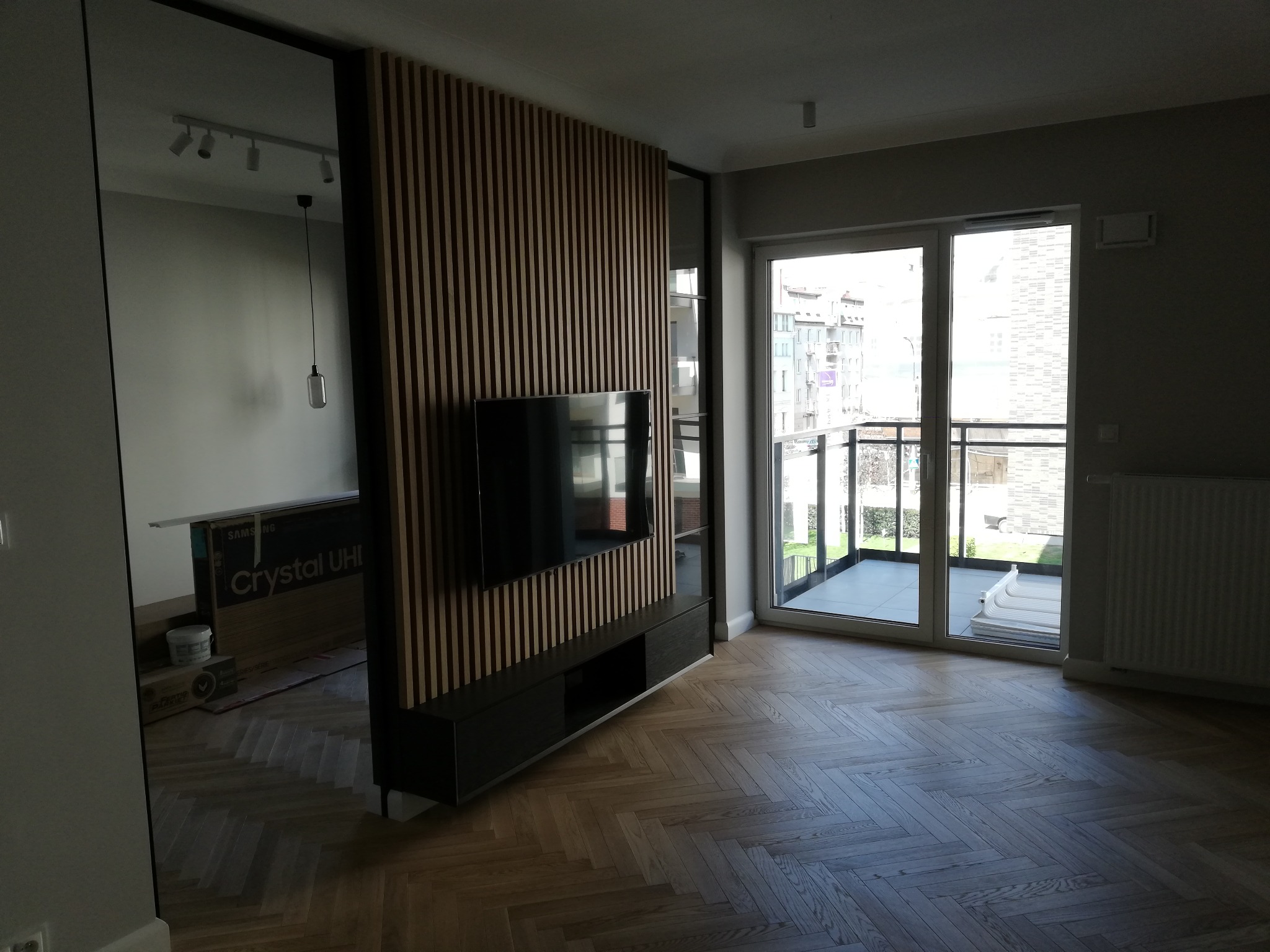 Salon po remoncie z drewnianą ścianką działową z telewizorem, parkiet ułożony w jodełkę, widok na balkon i rozpakowany telewizor w tle.