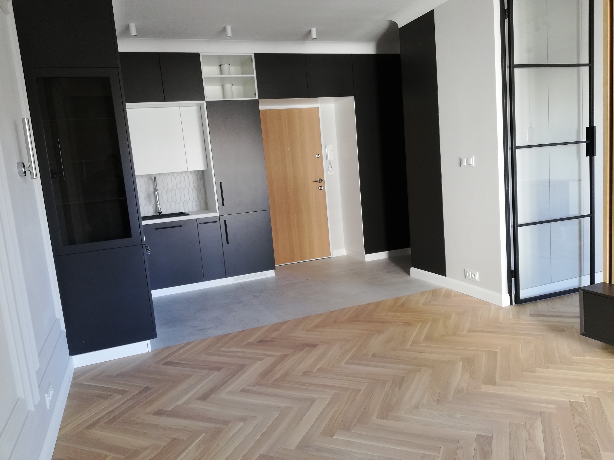 Nowoczesne wnętrze mieszkania w Warszawie z parkietem ułożonym w jodełkę, czarnymi meblami kuchennymi i przeszklonymi drzwiami w stylu loft.