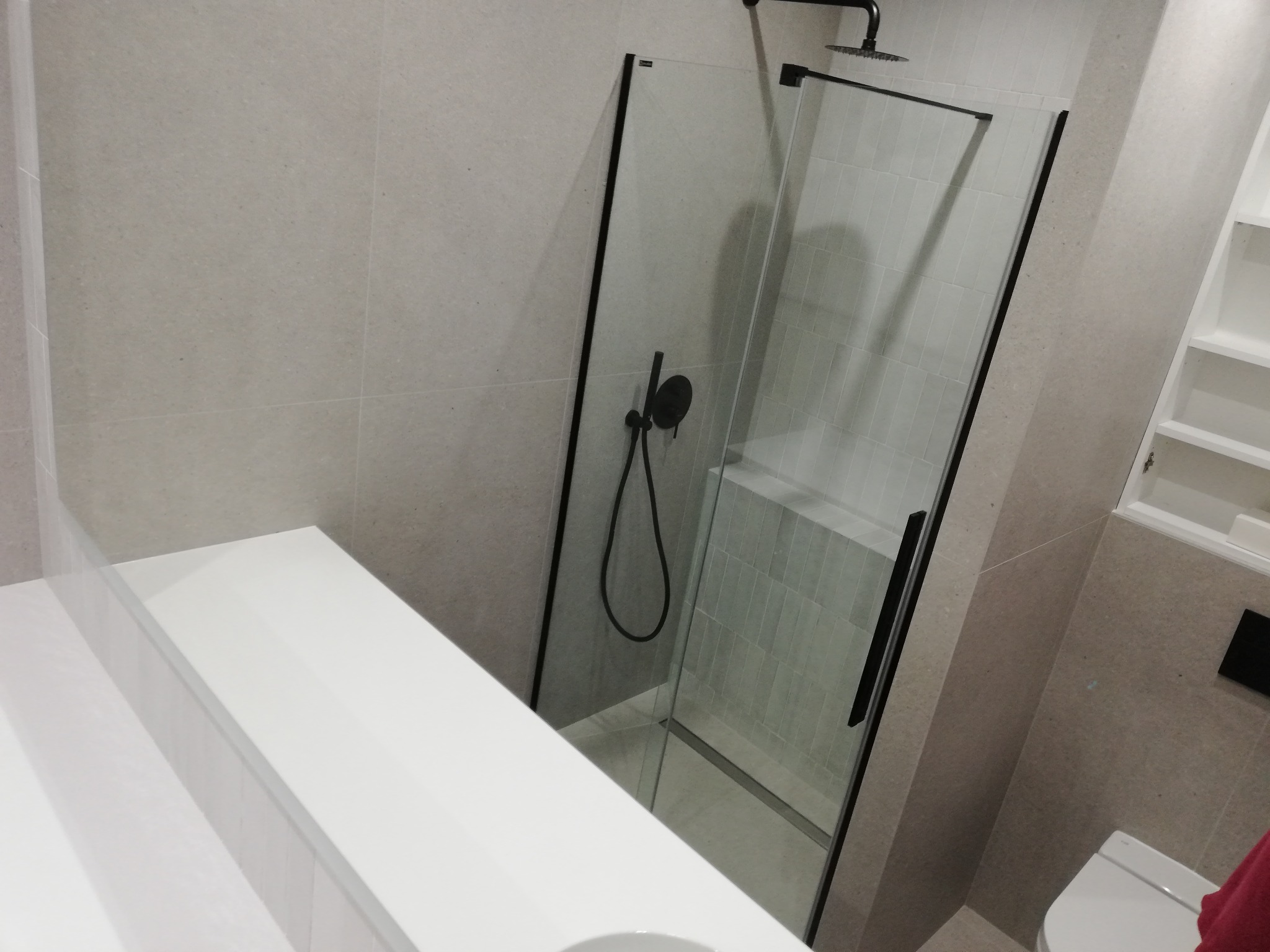 Nowoczesna kabina prysznicowa z czarnymi profilami i deszczownicą, jasne płytki imitujące kamień na ścianach, biała zabudowa umywalki z blatem, minimalistyczne wykończenie łazienki.