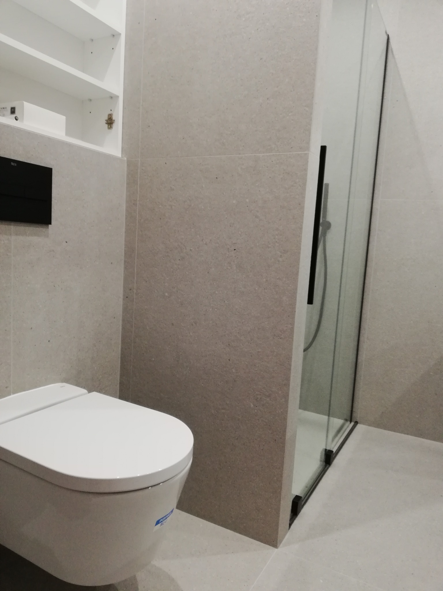 Nowoczesna łazienka z wiszącą toaletą, kabiną prysznicową ze szkła i szarymi płytkami na ścianach i podłodze.