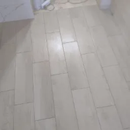 Ułożona podłoga z jasnych, drewnopodobnych płytek ceramicznych, widoczny worek z zaprawą i przedłużacz w tle.