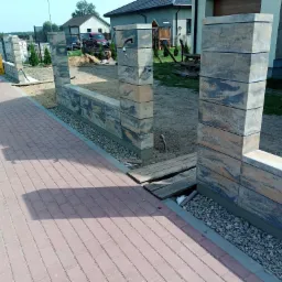 Wznoszenie murku z betonowych bloczków o nieregularnym wzorze, widoczny fragment chodnika z czerwonej kostki brukowej i przygotowany teren pod dalszą budowę, elementy instalacji wystające z muru.