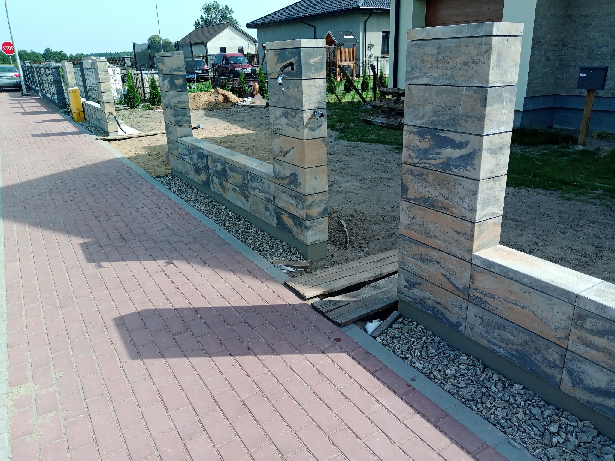 Wznoszenie murku z betonowych bloczków o nieregularnym wzorze, widoczny fragment chodnika z czerwonej kostki brukowej i przygotowany teren pod dalszą budowę, elementy instalacji wystające z muru.