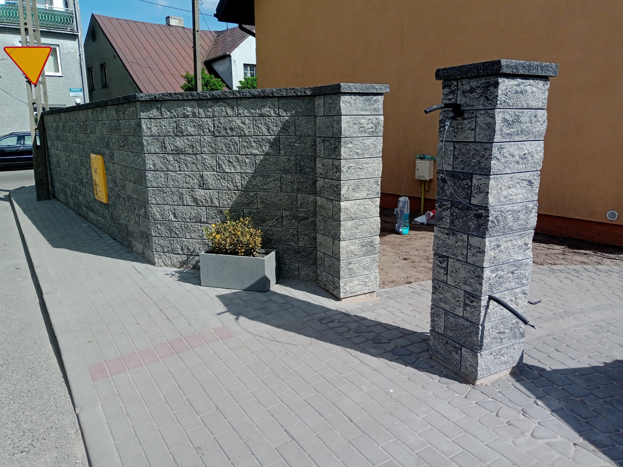 Nowoczesny mur z szarej kostki łupanej z donicą i słupkiem z kranem na chodniku z kostki brukowej, w tle budynki mieszkalne.