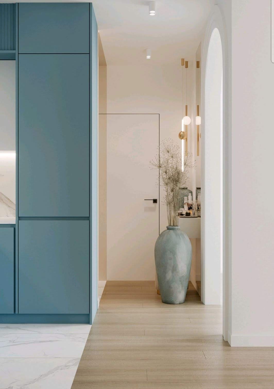 Nowoczesne wnętrze z błękitnymi szafkami kuchennymi, jasną drewnianą podłogą i dekoracyjnym wazonem z suszonymi kwiatami w przejściu do łazienki z oświetleniem punktowym.