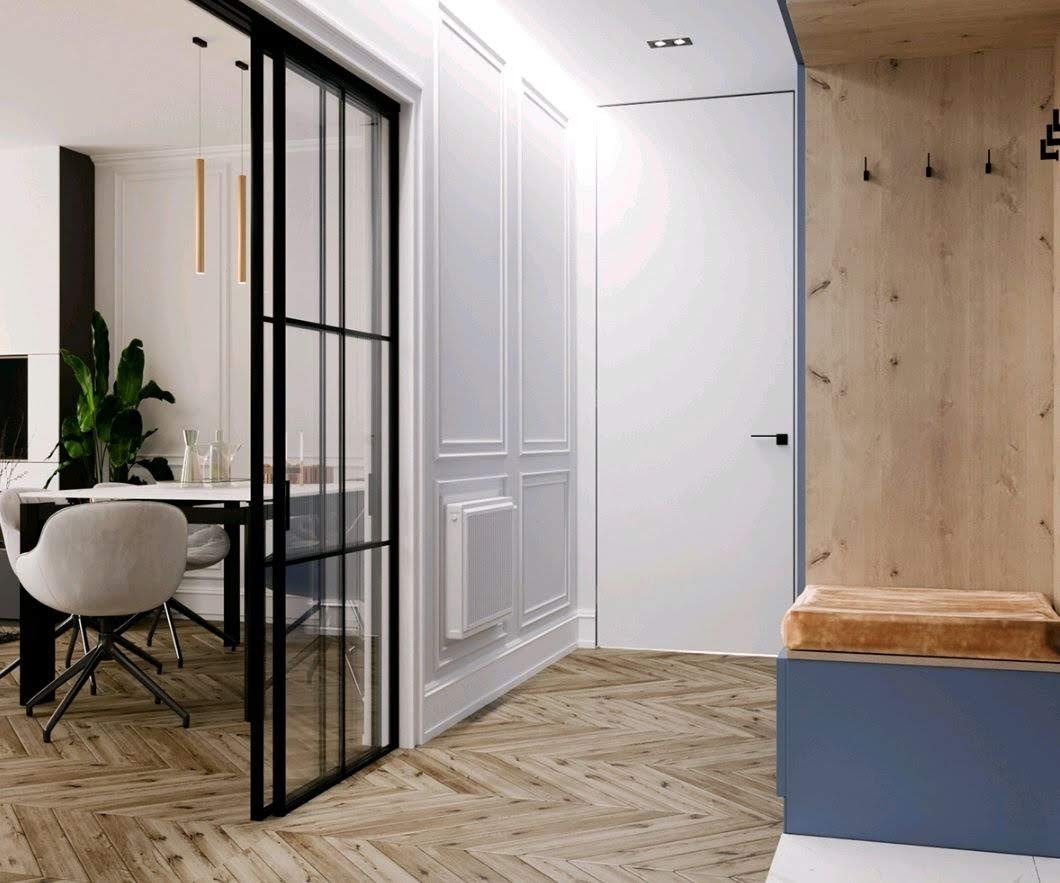 Nowoczesny przedpokój z drewnianą podłogą ułożoną w jodełkę, białe panele ścienne z dekoracyjnymi listwami, czarne przesuwne drzwi szklane i wbudowana szafka z siedziskiem w kolorze niebieskim...