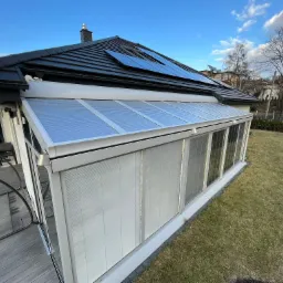 Zewnętrzny widok przeszklonej oranżerii z żaluzjami, dobudowanej do domu z czarnym dachem i panelami słonecznymi, w tle zielony trawnik i zabudowa miejska.