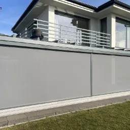 Zewnętrzne rolety przeciwsłoneczne w kolorze szarym, zamontowane na tarasie nowoczesnego domu z białymi ścianami i szarą balustradą. Widoczny fragment trawnika i błękitne niebo.