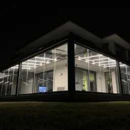 Nowoczesna, przeszklona oranżeria z balustradami szklanymi, oświetlona od wewnątrz, widoczna nocą z ogrodu.