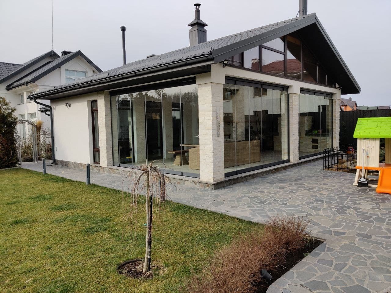 Nowoczesny dom z przeszkloną ścianą tarasową, wykorzystującą przesuwne panele szklane z czarnymi ramami, widoczne z zewnątrz na tle kamiennego chodnika i trawnika.