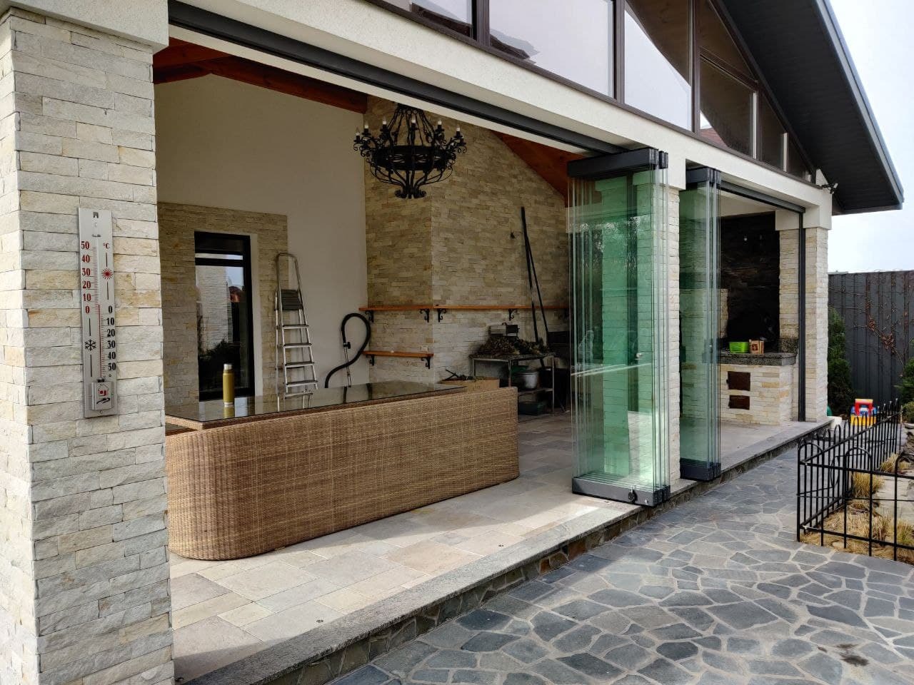 Zadaszony taras z rozsuwanymi szklanymi ścianami, widok na kamienną podłogę i wiklinowe meble, na ścianie termometr.