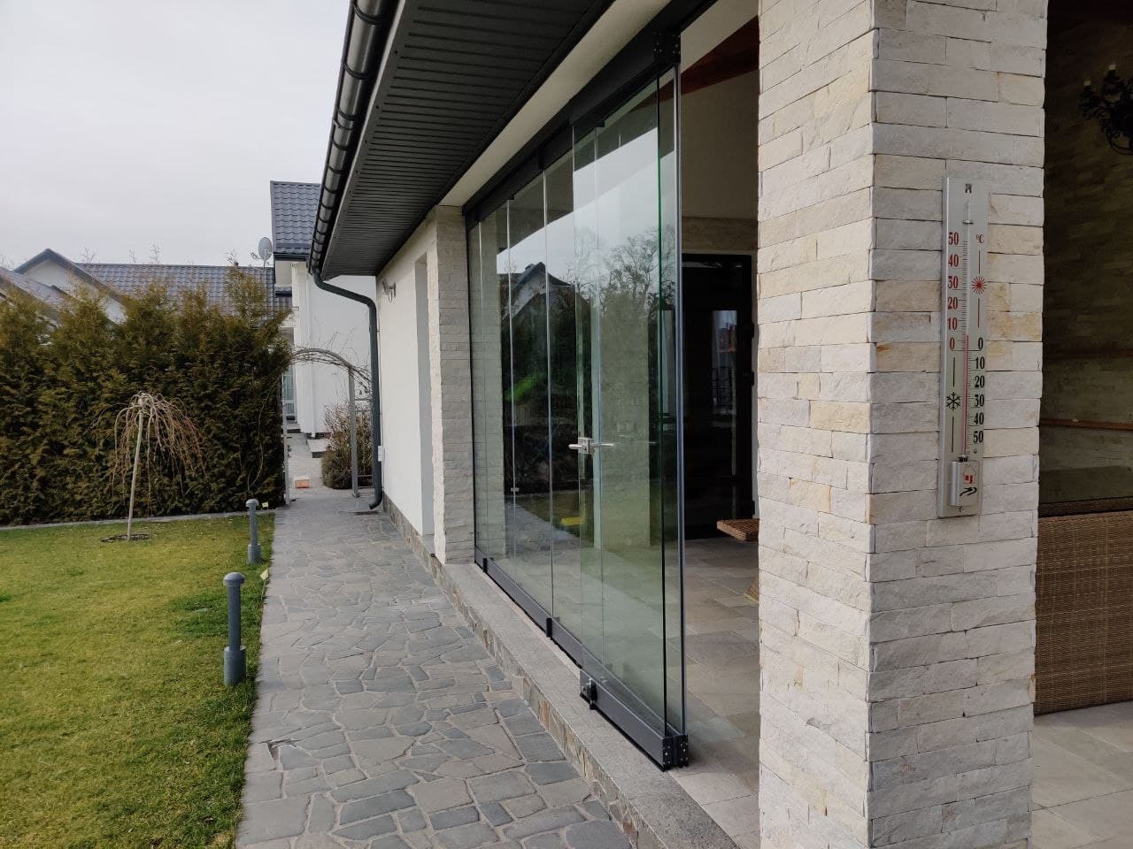Szklana zabudowa tarasu z przesuwnymi drzwiami, widok z zewnątrz na kamienną ścieżkę i zadbany trawnik, termometr na ścianie budynku.