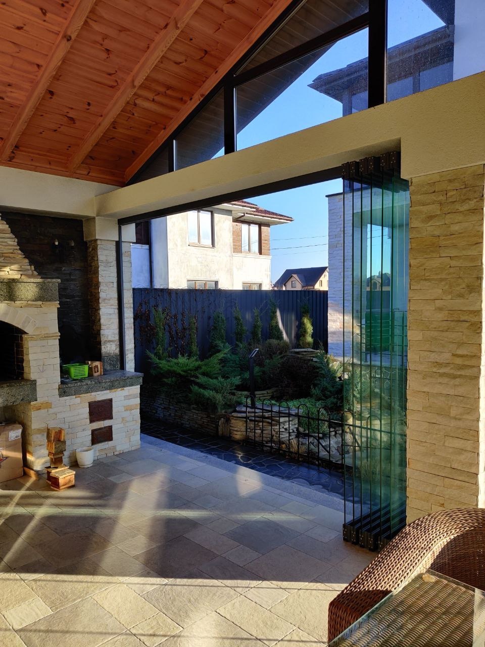 Zadaszony taras z kamienną podłogą i ścianą, wyposażony w składane szklane drzwi harmonijkowe, otwierające widok na ogród i dom.