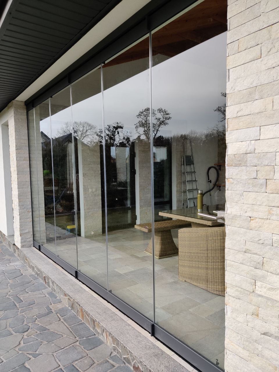 Szklana balustrada tarasowa z przesuwnymi panelami, widok z zewnątrz na kamienny taras i meble ogrodowe, minimalistyczna rama w kolorze antracytowym.