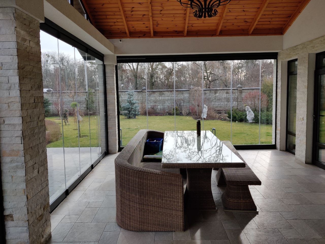 Szklana zabudowa tarasu z przesuwnymi panelami, widok na ogród ze statuami, rattanowe meble ogrodowe pod drewnianym sufitem z żyrandolem.