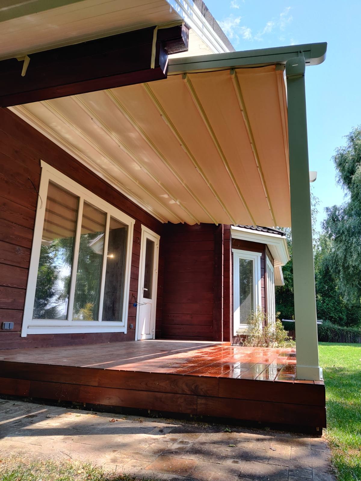 Zadaszony taras drewnianego domu z rozsuwaną markizą w beżowym kolorze, wspartą na białych kolumnach. Podłoga tarasu z polerowanego drewna, odbija światło słoneczne. Tło stanowi zielony trawnik...