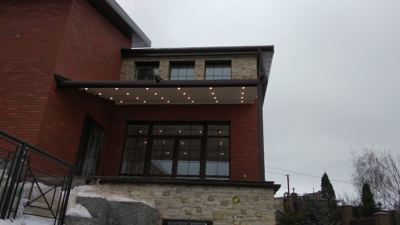 Zewnętrzny widok domu z cegły i kamienia z zamontowaną markizą z oświetleniem punktowym nad dużymi oknami.