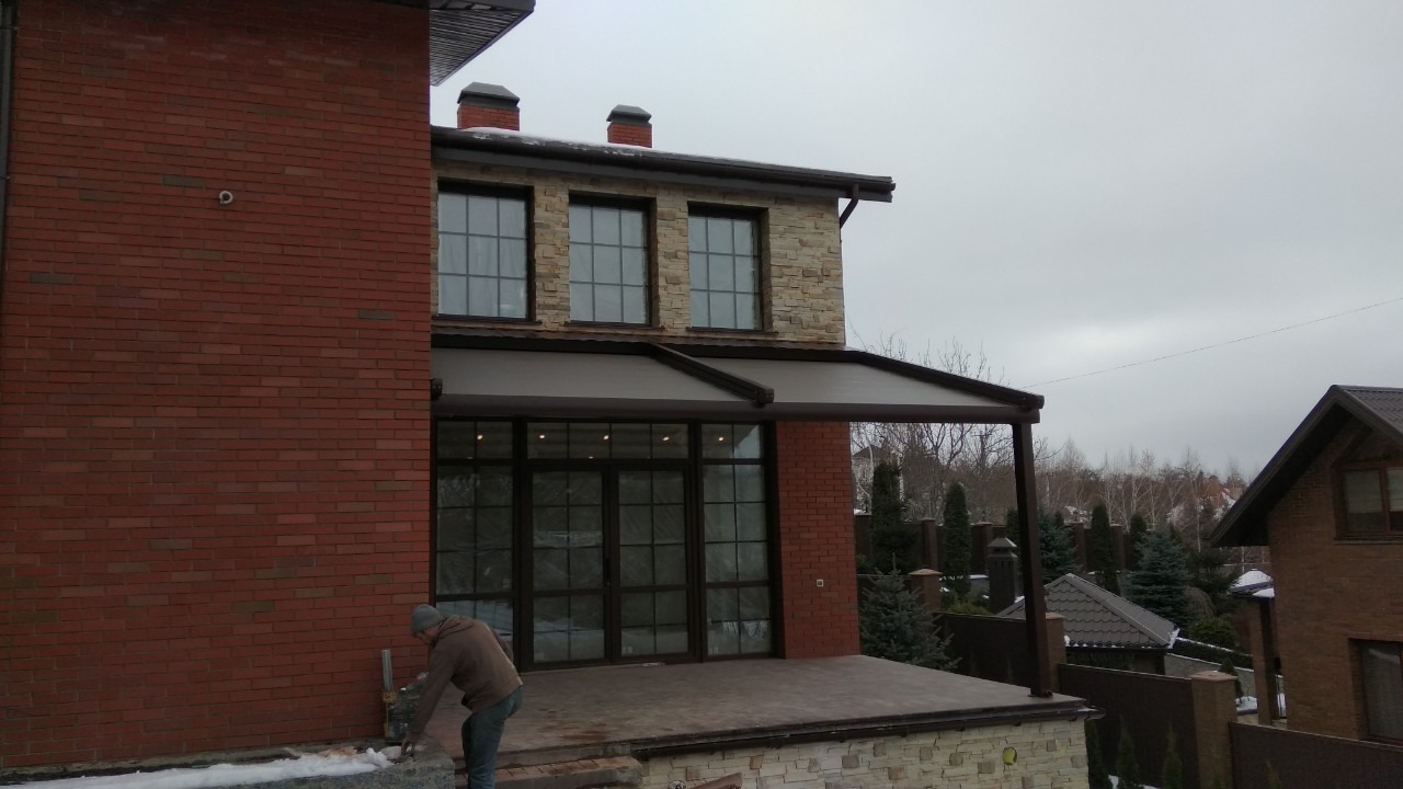 Zadaszony taras z dużymi szklanymi drzwiami i oknami w domu z cegły i kamienia, widoczna osoba pracująca przy wykończeniu.