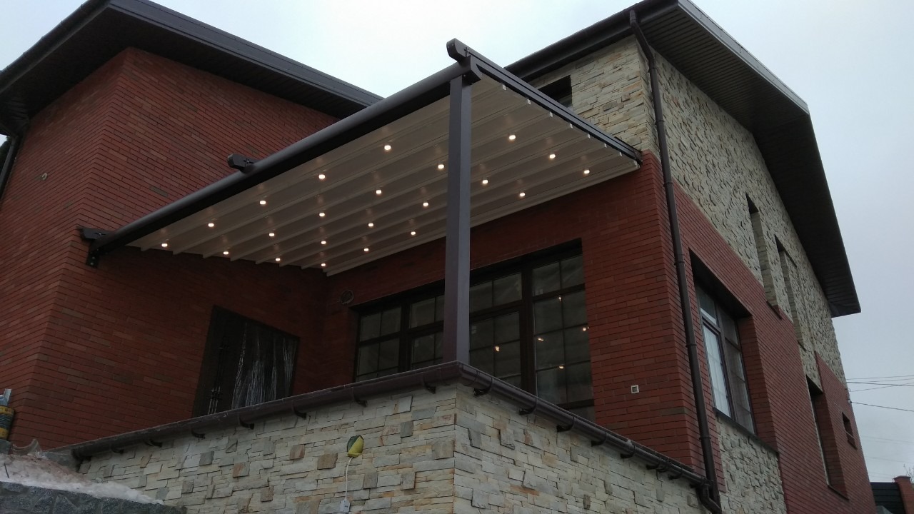 Zewnętrzny widok domu z cegły i kamienia z zamontowaną pergolą z oświetleniem punktowym, widoczne okna i rynny.