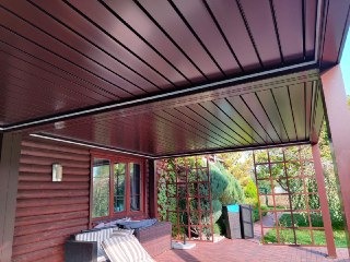 Zadaszenie tarasu z brązowych paneli, widok z poziomu tarasu na drewnianą elewację budynku i fragment ogrodu.