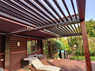 Zadaszony taras z regulowanymi lamelami, brązowa konstrukcja przylegająca do drewnianego domu, widoczny leżak na ceglanym patio i zieleń w tle.